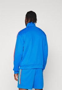 Giacca da tuta blu con collo alto e strisce laterali rosse, abbinata a pantaloni corti blu coordinati. Tessuto liscio con polsini e orlo elasticizzati.