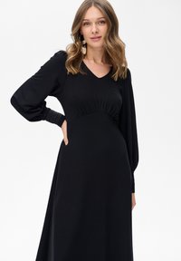 Kobieta z falującymi, jasnobrązowymi włosami ubrana w długą czarną suknię z bufkami i dużymi wzorzystymi kolczykami w kształcie kół, stojąca na białym tle.