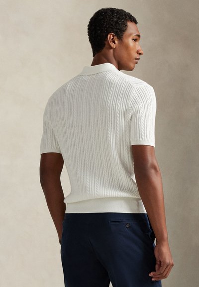 Homme de profil portant un polo en maille à manches courtes blanches avec des motifs torsadés et un pantalon bleu marine.