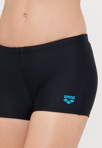 Shorts de bain noirs en tissu extensible, présentant un design ajusté et une taille élastique. Le logo "arena" est affiché en bleu.