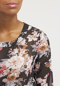Blusa nera a fiori con scollo rotondo, caratterizzata da grandi fiori rosa e beige, tessuto trasparente e delicate fantasie botaniche.