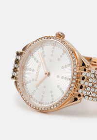 Złoty zegarek w kolorze różowego złota z bransoletką wysadzaną kryształami, tarczą w białym kolorze o efektach słonecznych oraz zapięciem ozdobionym błyszczącymi kamieniami. Regulowana korona.
