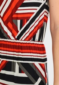 Klädets midja med ett geometriskt randigt mönster i rött, svart, vitt och grått samt ett elastiskt cinchband, höger arm synlig mot ljus bakgrund.