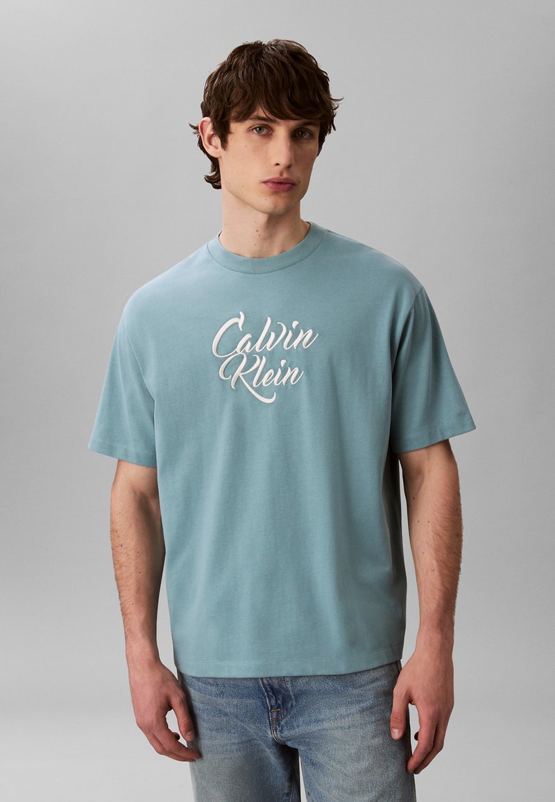 Jeune homme portant un t-shirt Calvin Klein bleu clair et un jean bleu délavé, debout devant un fond gris uni.