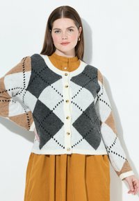 Žena oblečená v zapínacím argyle svetříku v bílé, šedé a béžové barvě přes hořčicové šaty, stojící s jednou rukou na boku.