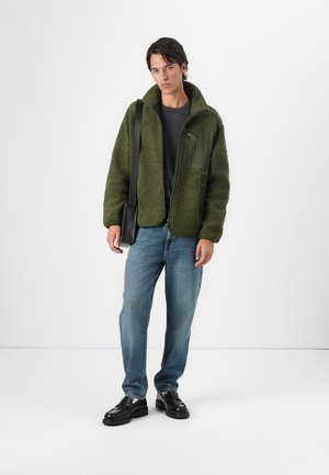 Chaqueta de forro polar verde con un bolsillo en el pecho con cremallera, llevada sobre una camisa oscura, combinada con jeans azules y mocasines negros. Diseño texturizado y casual.