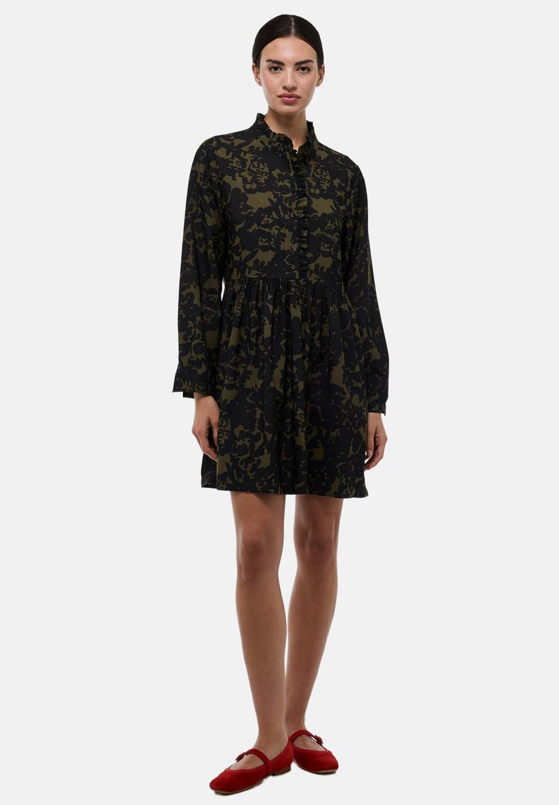 Robe à manches longues de couleur vert foncé avec un motif floral noir, dotée d'un col à volants, d'une patte de boutonnage et d'une silhouette en A. Portée avec des ballerines rouges.