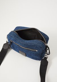 Dunkelblaue Denim-Umhängetasche mit einem oberen Reißverschluss, schwarzem Beschlag und verstellbarem Gurt. Das Innere ist mit einem schlichten schwarzen Stofffutter ausgestattet.