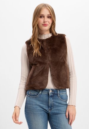 Giovane donna con lunghi capelli biondi che indossa un gilet in pelliccia sintetica marrone sopra un dolcevita beige a coste e jeans blu su sfondo bianco.