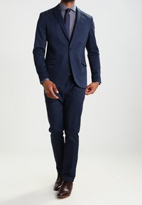 Marinblå kostym med skräddarsydd passform, funktionell slagkrage, en-knapplikning och byxor med rak ben. Kombinerad med en ljusblå skjorta och slips.