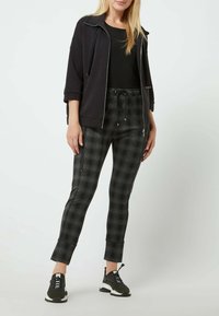 Svart zip-up-jacka över en svart topp, i kombination med skräddarsydda rutiga byxor i grå nyanser. Modellen bär svarta sneakers med vita accenter.