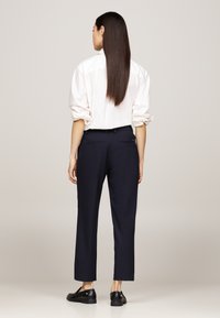 Camicia bianca con le maniche arrotolate, abbinata a pantaloni scuri blu navy e scarpe nere. I pantaloni hanno una vestibilità aderente e dettagli minimi.