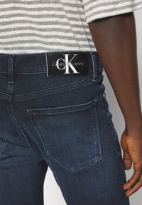 Jeans de color azul oscuro con una etiqueta negra de Calvin Klein Jeans en la cintura, combinados con una camiseta de rayas en gris y blanco y un brazo derecho visible.