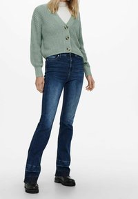 Cardigan texturizado en verde menta con tres botones, combinado con jeans acampanados oscuros y botas negras gruesas. Diseño y ajuste minimalista.