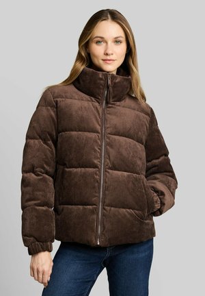 Junge Frau trägt eine braune wattierte Cordjacke mit Reißverschluss und hohem Kragen, steht mit einer Hand in der Tasche vor grauem Hintergrund.