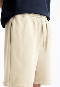 Gros plan sur un short beige clair avec cordon de serrage, porté avec une chemise bleu marine foncé, montrant des poches latérales et une taille élastique.