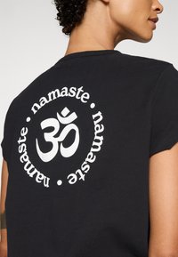 Crna pamučna majica s kružnim bijelim dizajnom s simbolom Om i tekstom "namaste" istaknutim na leđima.