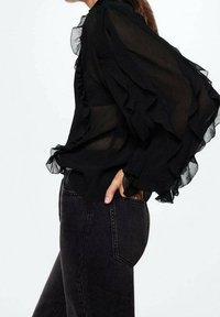 Blusa nera trasparente con maniche a volant, caratterizzata da un colletto alto e una texture morbida, abbinata a jeans neri a vita alta.