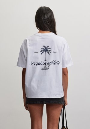 Femme aux longs cheveux foncés portant un T-shirt blanc avec un graphisme de palmier et le texte « Pegador athletic club », tenant un sac noir derrière.