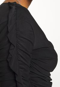 Svart topp med tygvolanger längs axeln, med en texturerad, samlad design som skapar en figurnära silhuett.