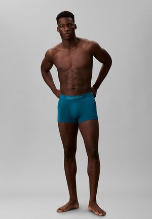 Muskularny mężczyzna stoi boso, z rękami za plecami, ubrany w turkusowe bokserki Calvin Klein na jednolitym szarym tle.