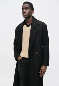 Fekete gyapjú kabát bemetszett ággall, bézs kötött V-nyakú pulóverrel alatta, és sötét farmernadrággal. Gombbal és oldalzsebekkel rendelkezik.