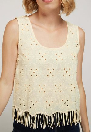 Femme portant un haut sans manches en dentelle crème avec des motifs floraux découpés et un ourlet à franges, associée à un pantalon foncé.