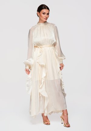 Vrouw met een lange, vloeiende satijnen jurk in crèmewit met geplooide halslijn, gestrikte taille, gerafelde zoom en doorschijnende lange mouwen, staand met gouden hakken.