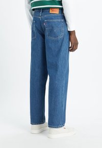 Persona con jeans de denim azul de corte holgado y zapatillas blancas, vista desde atrás sobre un fondo blanco uniforme.