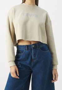Felpa corta beige con logo "LEVI'S" in grigio chiaro, caratterizzata da maniche lunghe e scollatura rotonda. Abbinata a jeans in denim blu a vita alta.
