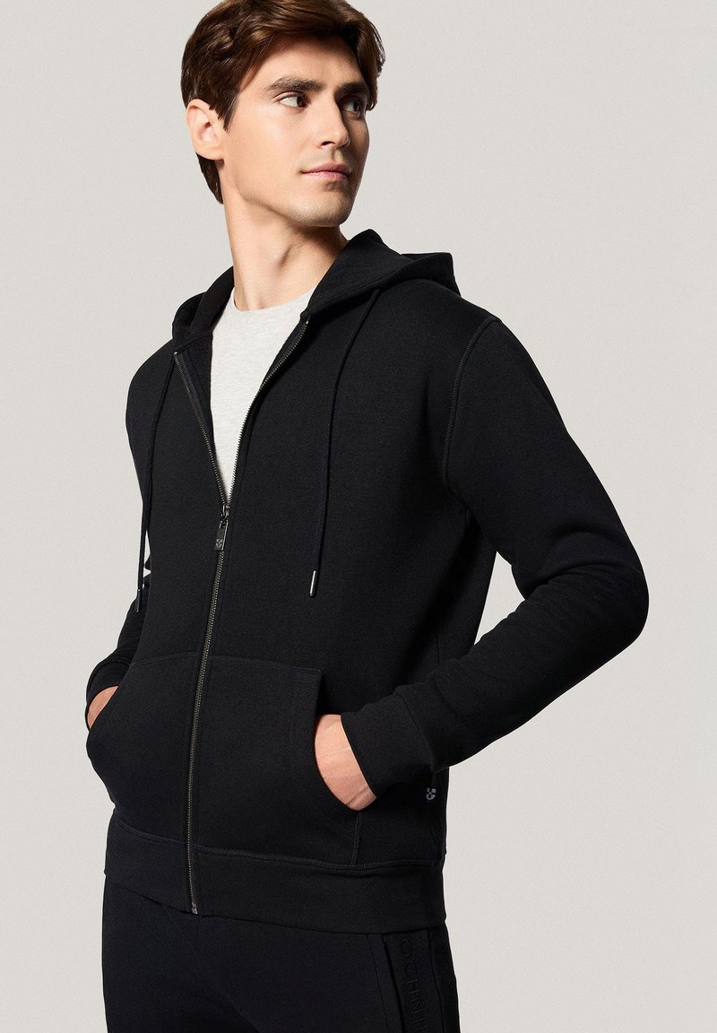 Czarna bluza z kapturem na zamek, wykonana z miękkiego materiału, z przednią kieszenią i sznurkami do regulacji. Model stoi z rękami w kieszeniach.