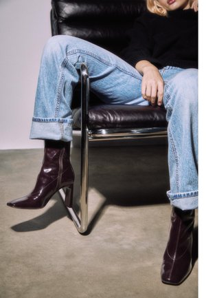 Viininpunaiset patenttinahkaiset nilkkurit, joissa on terävä kärki ja paksu korko; yhdistetty vaaleansinisiin, käännettyihin farkkuihin, istuen kromisella tuolilla.