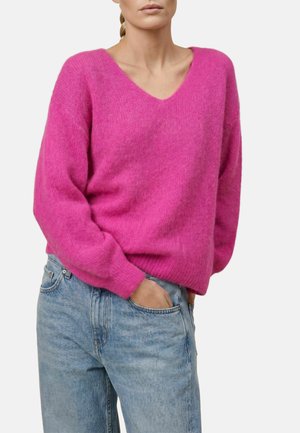 Femme portant un pull rose vif à col en V et un jean bleu clair, debout avec une main dans la poche sur un fond uni.
