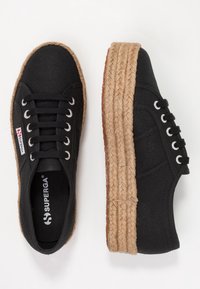 Zapatillas de lona negras con suela de yute trenzada y elevada. Presentan ojales plateados y una etiqueta de logo en el lateral. Diseño de puntera redonda.