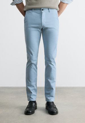 Mand iført lyseblå slim-fit bukser, sorte loafers og en beige sweater med opkrullede, stribede ærmer, stående på en grå gulv.