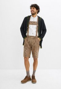 Lederhosen in suede marrone con ricami dettagliati, abbinati a una camicia bianca con bottoni e un cardigan scuro con accenti verdi. Scarpe marroni e calze grigie.