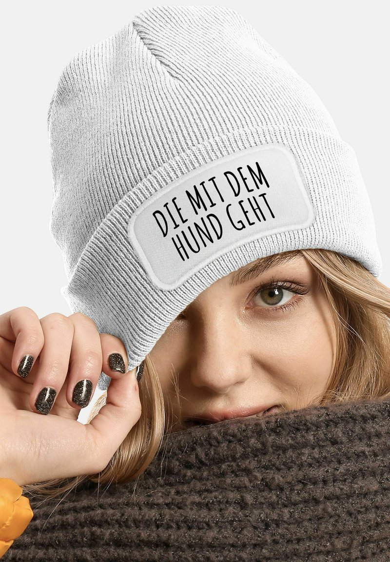 White knit beanie with a rectangular patch displaying the text "DIE MIT DEM HUND GEHT." Model's hand is resting on a brown knitted scarf.