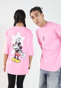 Rosa oversized t-skjorte med et svart og hvitt Mickey Mouse-trykk på baksiden, med en mindre versjon på forsiden. Stjernesformet detalj.