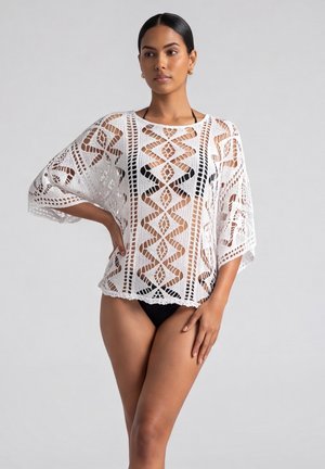 Vrouw draagt een wit gehaakt overslagstuk over een zwarte bikini, staat met één hand op haar heup tegen een effen lichte achtergrond.