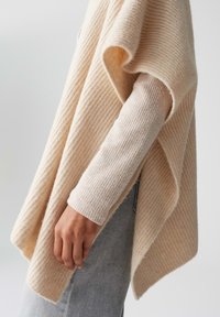 Beige gestrickter Poncho mit gerippter Textur, lockerer Passform und asymmetrischem Fall. Über einem hellen, gerippten Langarmoberteil getragen.