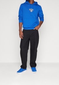 Blå huvtröja med en framficka, snörning och logotyp. Kombinerad med svarta, löst sittande byxor och blå sneakers med vita sulor.