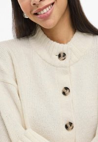 Kremfarget strikket cardigan med høy krage, ribbestrikket detaljering og store, runde knapper med marmormønster.