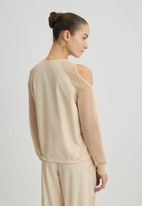 Beige sporttop met lange mesh-mouwen en uitsnijding bij de schouders. Beschikt over een ronde hals en een elastische zoom voor een getailleerde look.