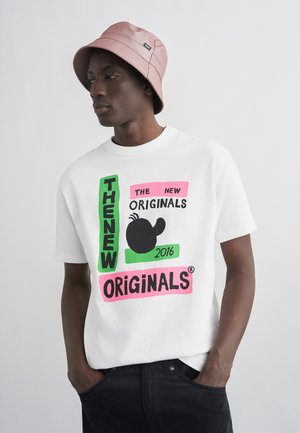 Mann som bærer en lyserosa bøttehatt, hvit t-skjorte med fargerik "The New Originals 2016"-tekst og grafikk, og svarte bukser, med hendene i lommene.