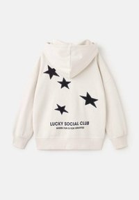 Hoodie crème avec une capuche doublée. Présente des appliqués en forme d'étoiles noires et le texte "LUCKY SOCIAL CLUB WHERE FUN IS FOR GRANTED" au dos.