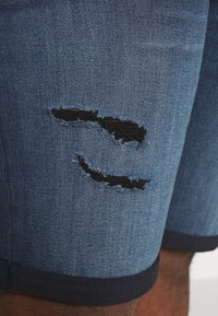 Shorts de mezclilla con una sección desgastada y un parche negro. Tela azul, dobladillo enrollado y una textura suave.
