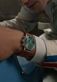 Chronograph-Uhr mit grünem Ziffernblatt, silbernem Gehäuse und braunem Lederarmband. Verfügt über drei Hilfszifferblätter und eine Datumsanzeige auf 6 Uhr.
