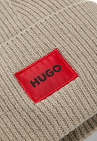 Bonnet en tricot beige avec un motif texturé. Présente une étiquette rectangulaire rouge avec "HUGO" en noir. Matériau en fil doux et épais.