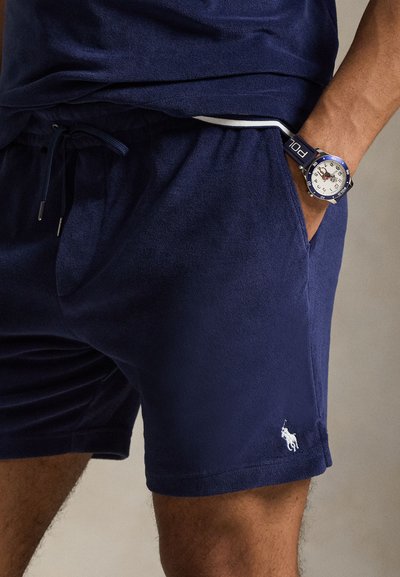 Σκούρα μπλε σορτς από απαλό ύφασμα με ελαστική μέση και κορδόνι, με ένα μικρό λευκό λογότυπο στο τελείωμα. Μπλε ρολόι με λευκό καντράν.