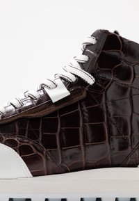 Sneaker montante en cuir embossé crocodile marron foncé avec des lacets blancs. Présente une semelle en caoutchouc blanche et des accents noirs. Texture lisse.